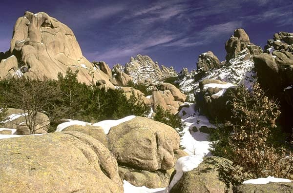 Large rocky formations with patches of snow under a clear blue sky, surrounded by sparse vegetation in a mountainous area