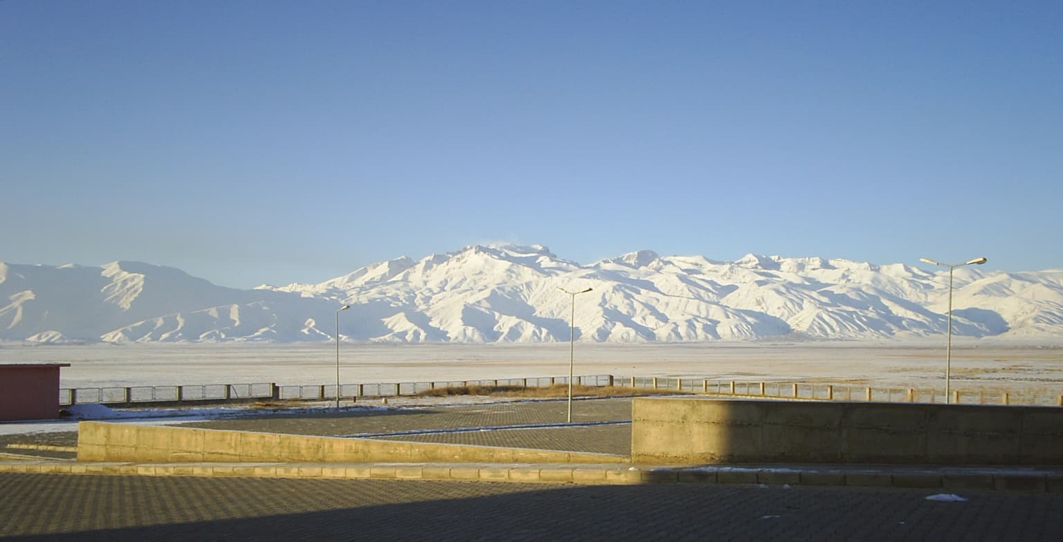 Wide landscape view of snow-covered mountain peaks under clear blue sky with a concrete structure and paved area in the foreground