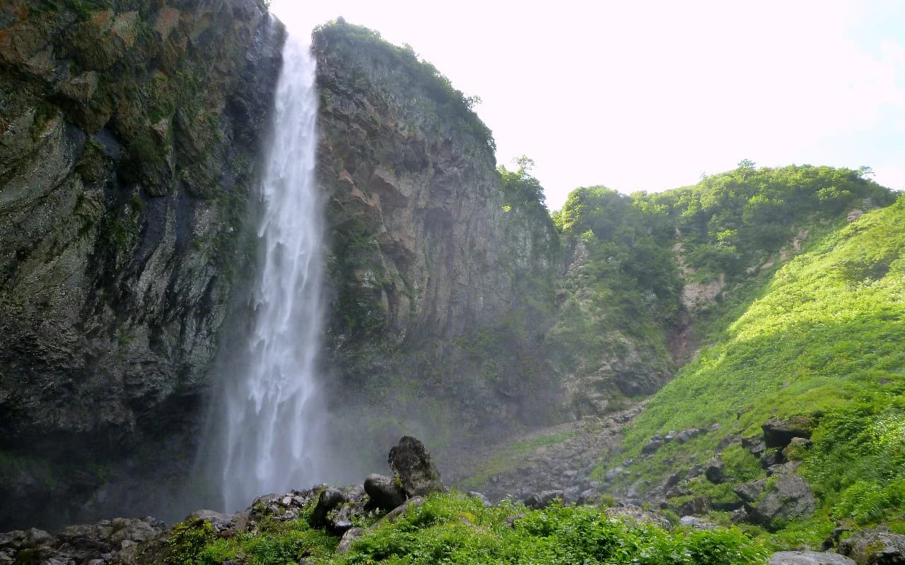 Cascada alta cayendo por un acantilado rocoso con vegetación verde exuberante en primer plano y fondo