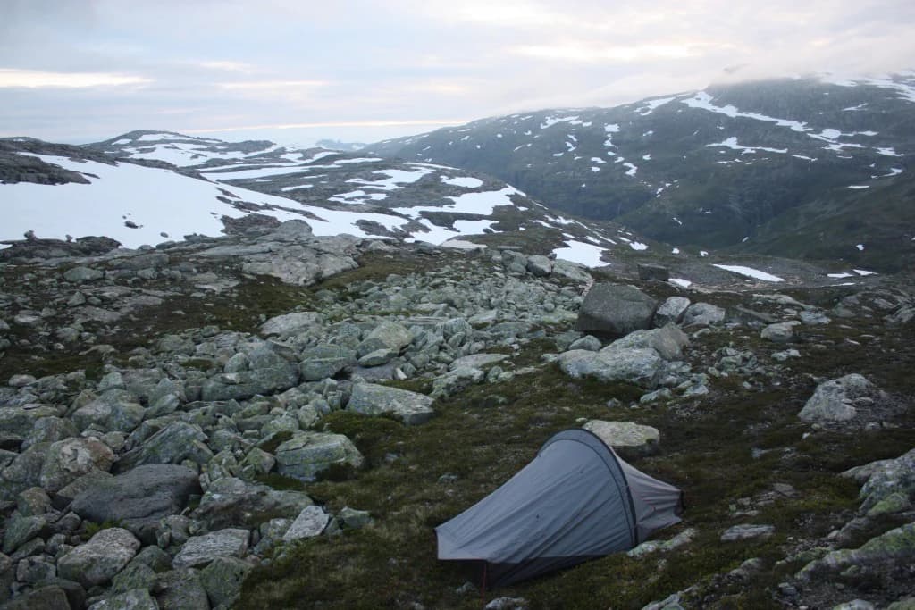 Gray hiking tent on rocky ground with patches of snow and distant mountain peaks under an overcast sky