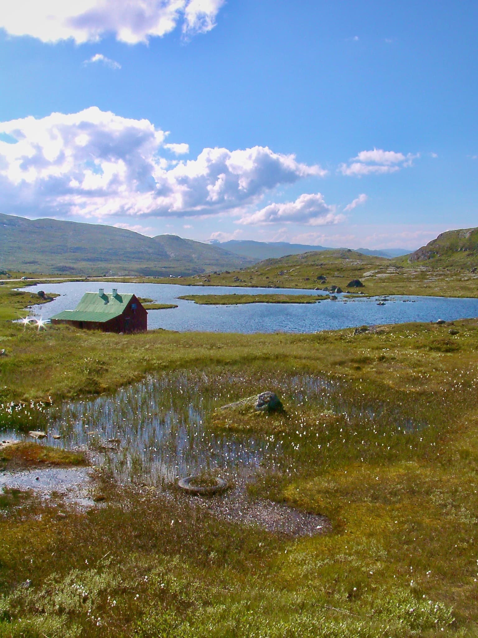 Landscape of Hardangervidda National Park featuring a small red cabin with green roof, a lake, grassy terrain with scattered water pools, and distant mountains under a blue sky with white clouds