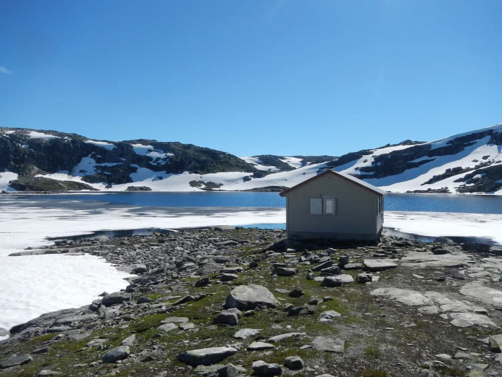 A small gray cabin on rocky terrain with snow patches, a lake, and snow-capped mountains under a clear blue sky