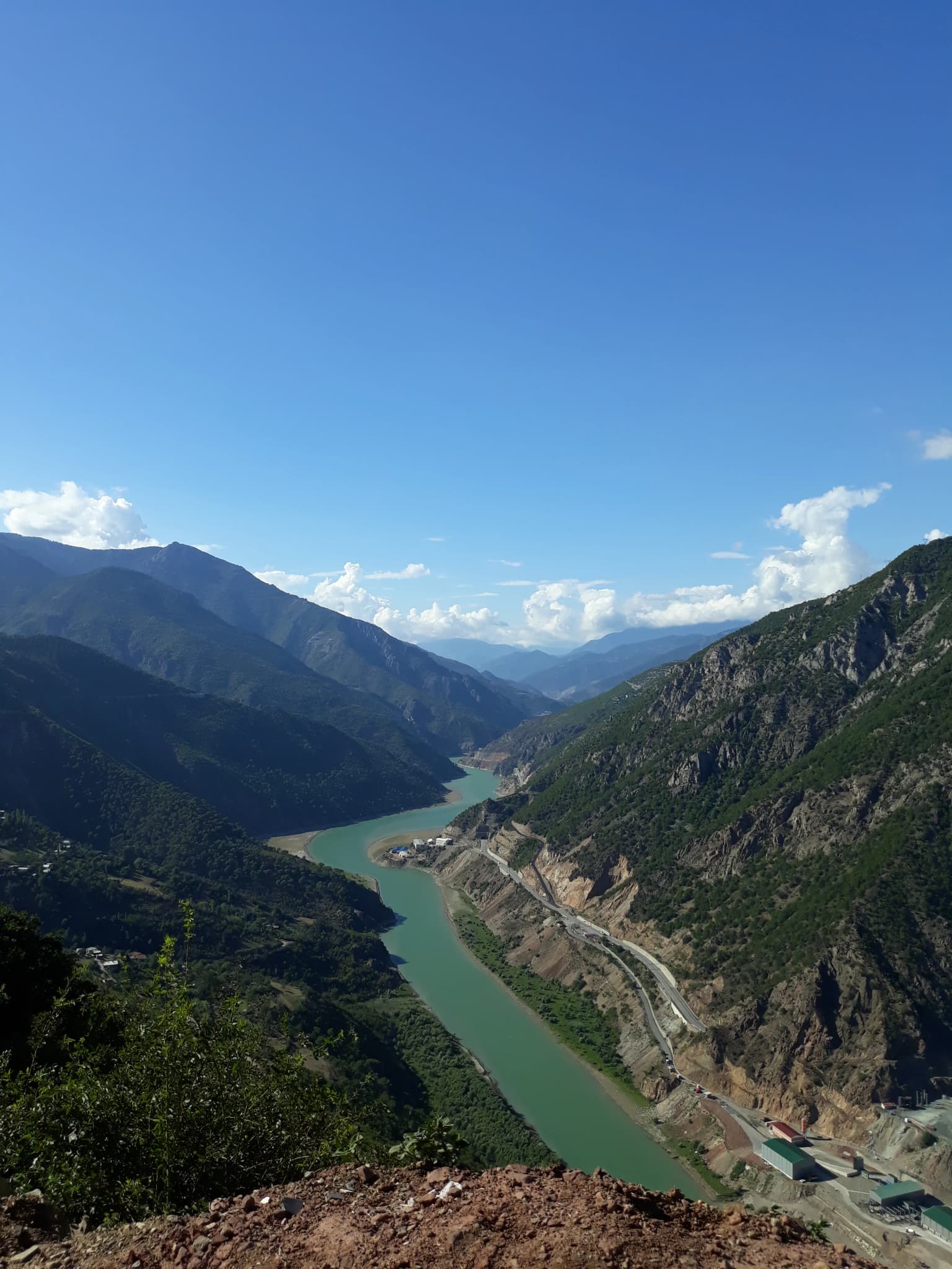 Wide-angle view of a turquoise river flowing through a mountainous valley with steep rocky cliffs and lush green vegetation under a clear blue sky.
