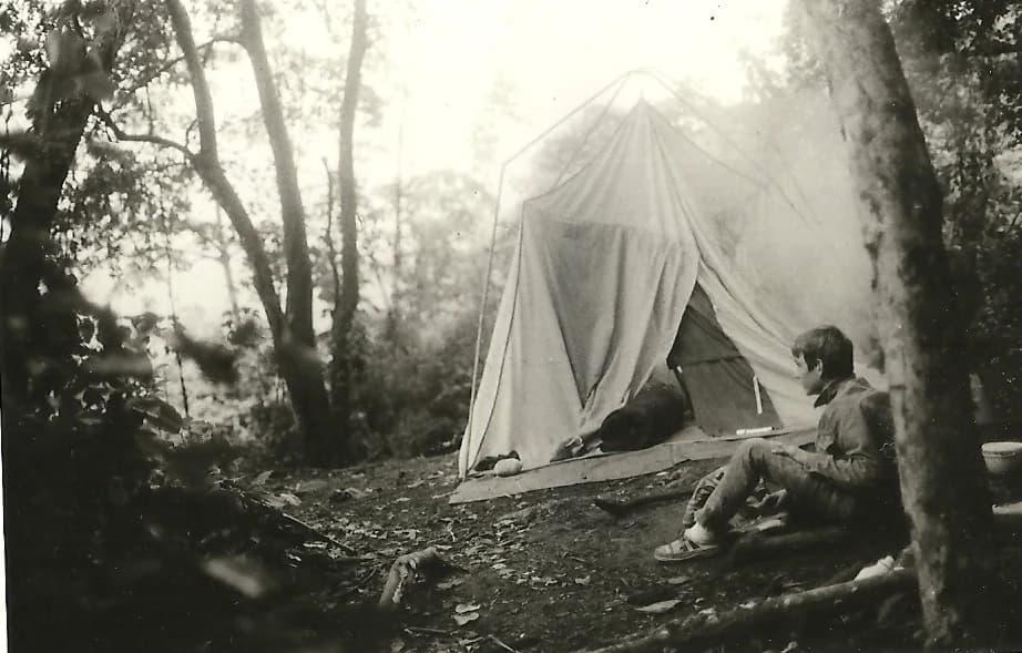 Black-and-white photograph of a person sitting next to a canvas tent in a forested area with trees and foliage surrounding the campsite