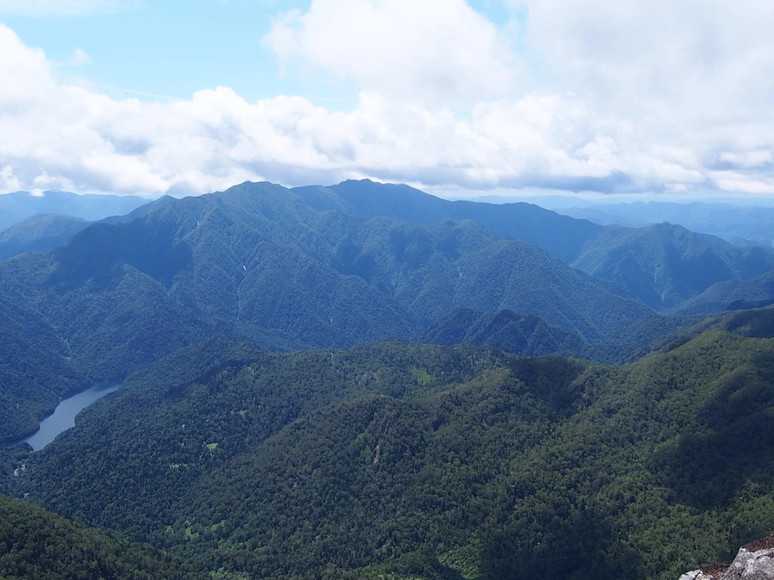 Wide landscape view showing mountain ranges, forested valleys, and a lake under a partly cloudy sky