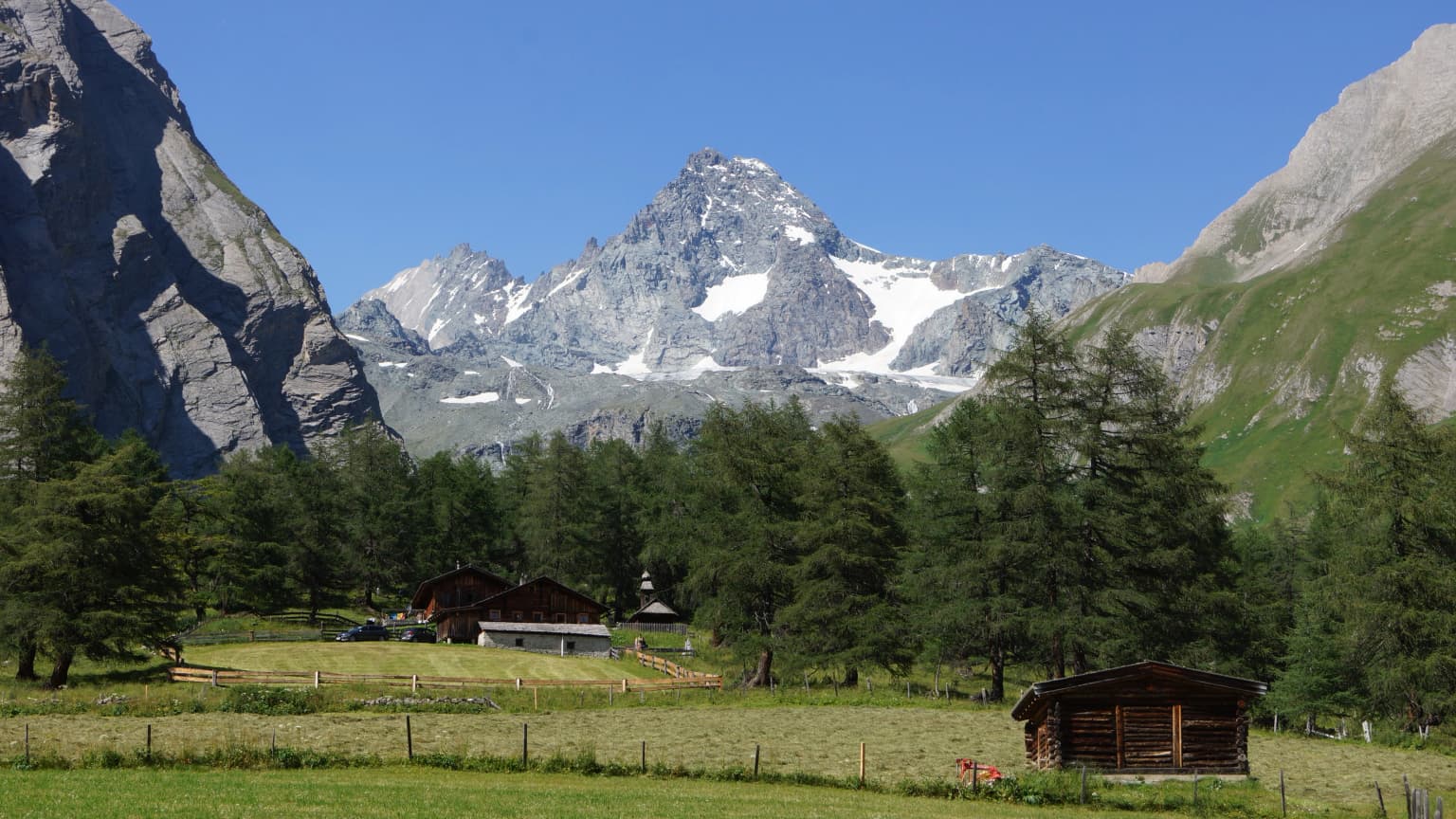 Alpine landscape with mountain valley, wooden huts, and snow-capped mountain peak under clear blue sky