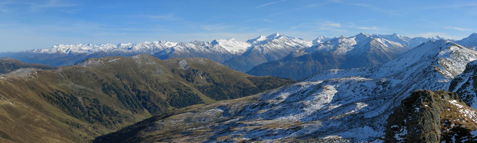 Wide landscape of snow-capped mountains, valleys, and rocky slopes under a clear blue sky