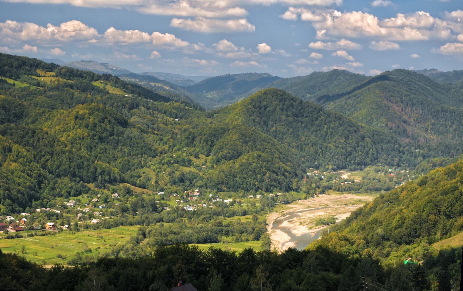 Mountainous landscape with green valleys, a river, and distant hills under a partly cloudy sky