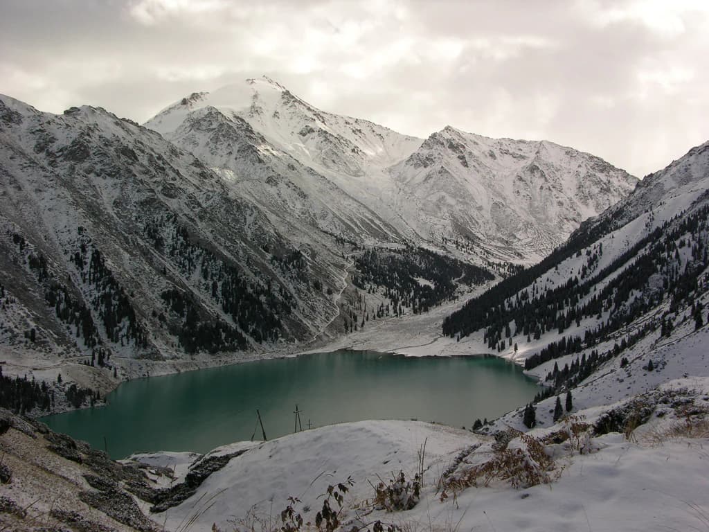 Snow-covered mountain range with a turquoise lake in a valley under an overcast sky