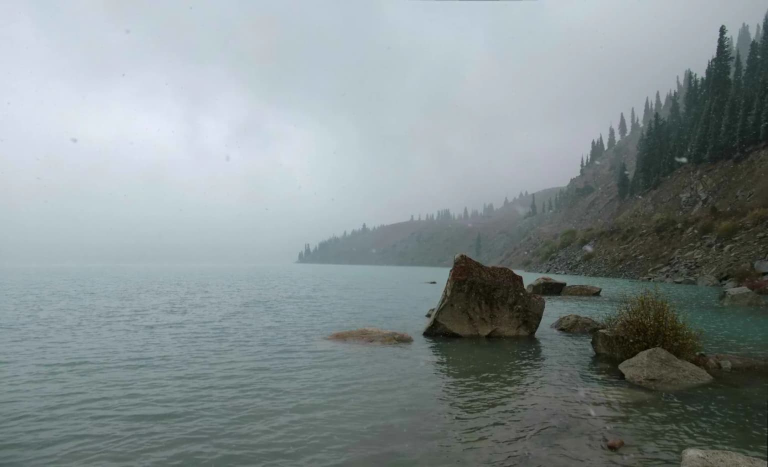 Calm lake water with large rocks in the foreground, a forested mountain slope in the background, and foggy sky