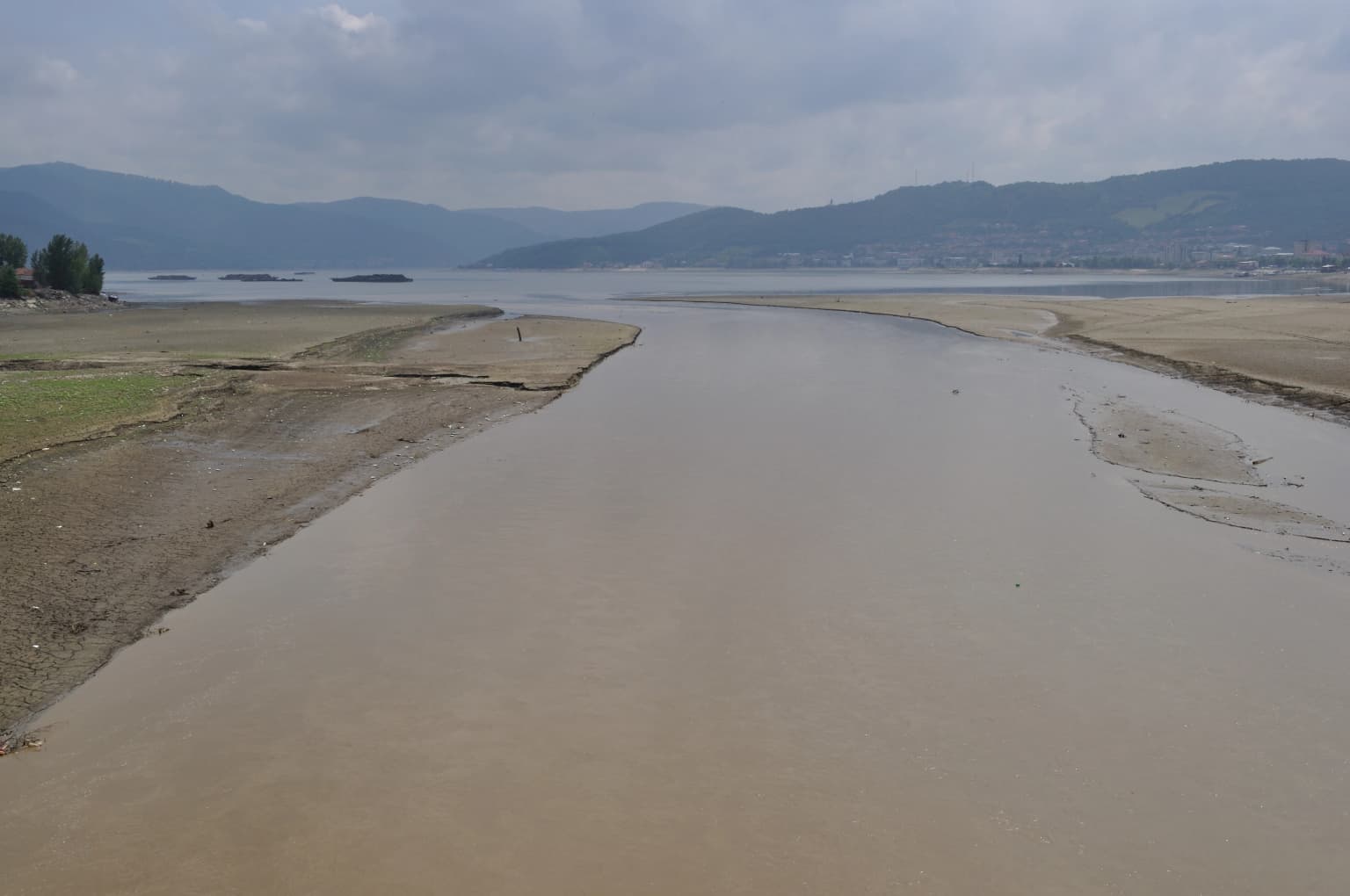 Muddy river flowing into a wider waterway with grassy banks and mountains in the distance