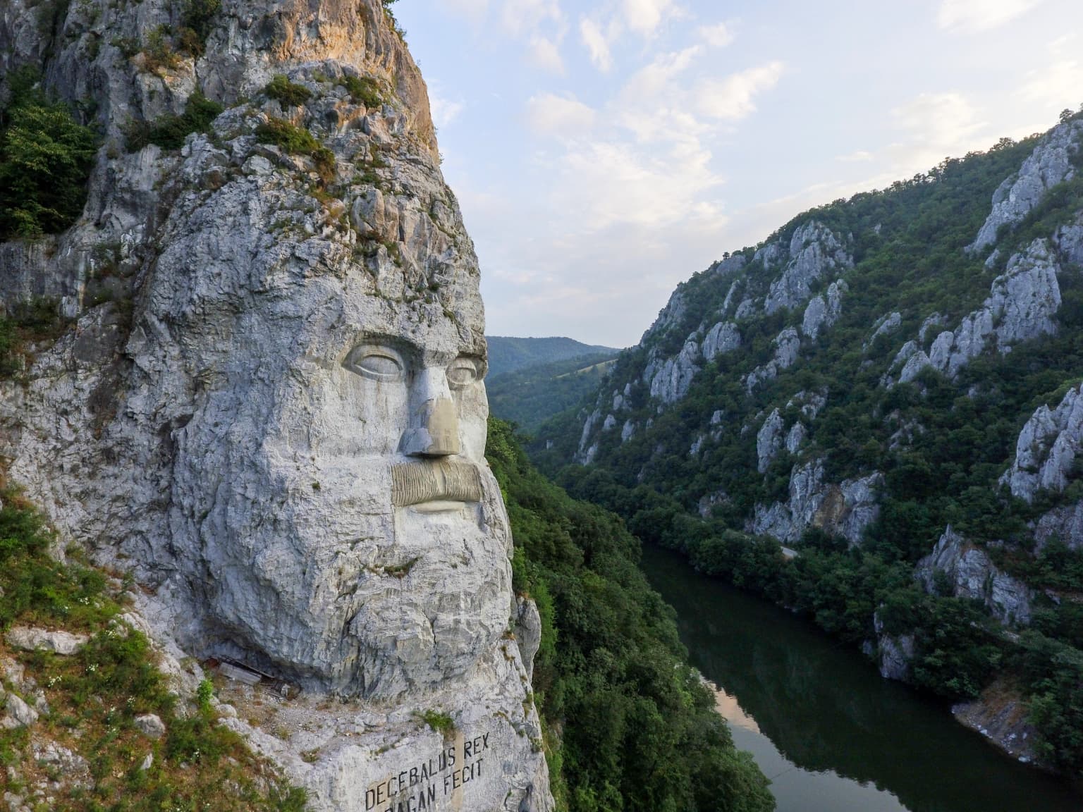 Large stone face carving on a cliffside with a river and mountainous landscape below