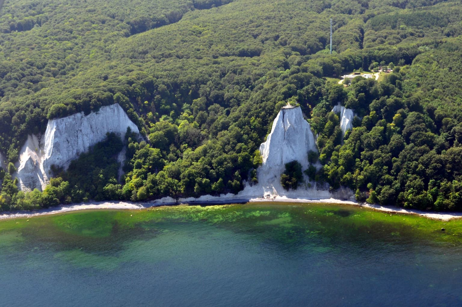 Aerial view of white chalk cliffs, dense green forest, and turquoise water along the coastline