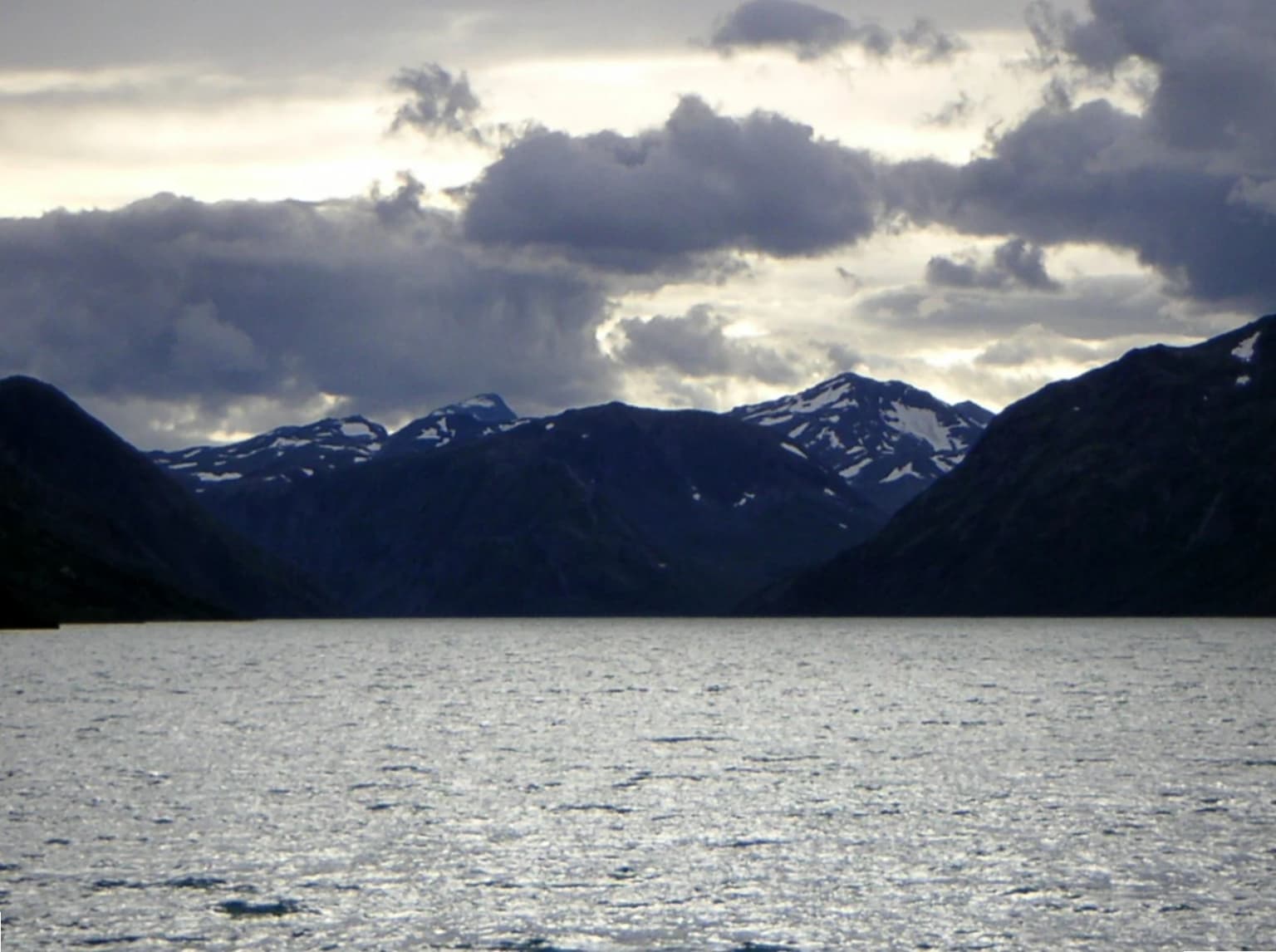 A lake with calm water reflecting light under a cloudy sky, surrounded by dark mountains with snow patches