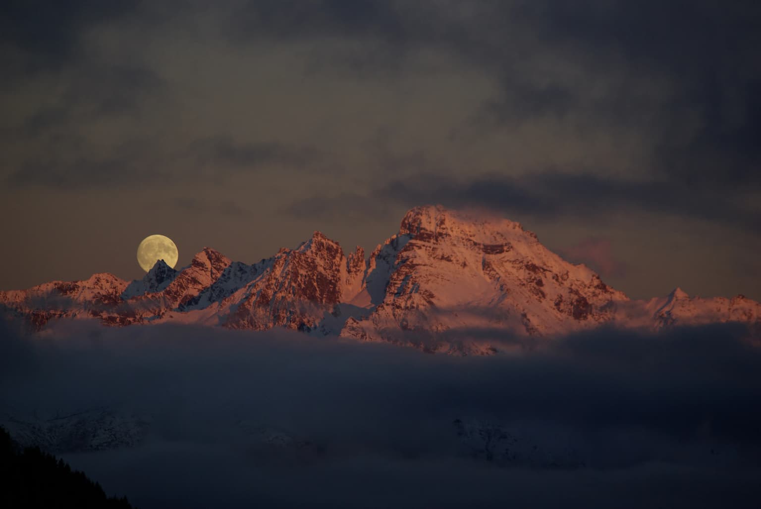 Snow-covered mountain peaks under twilight sky with full moon visible above mountains and misty clouds below