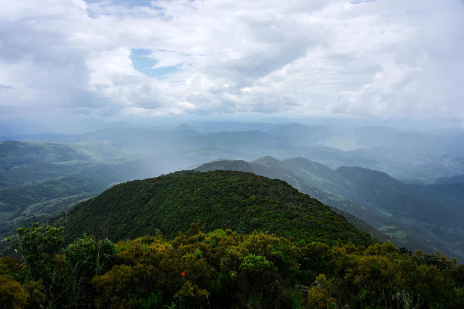 Wide landscape view of Mount Kahuzi summit with dense forested slopes, rolling mountains in the background, and partly cloudy sky