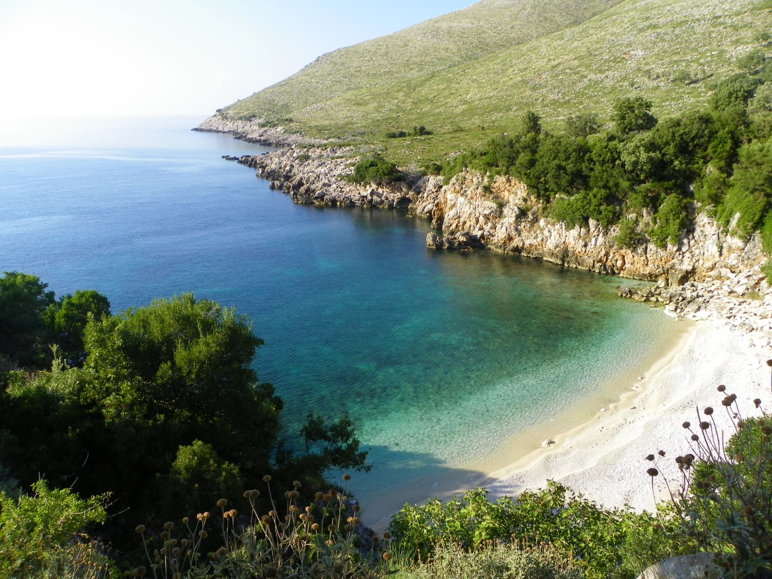Coastal bay with clear turquoise water, sandy beach, rocky cliffs, and green vegetation