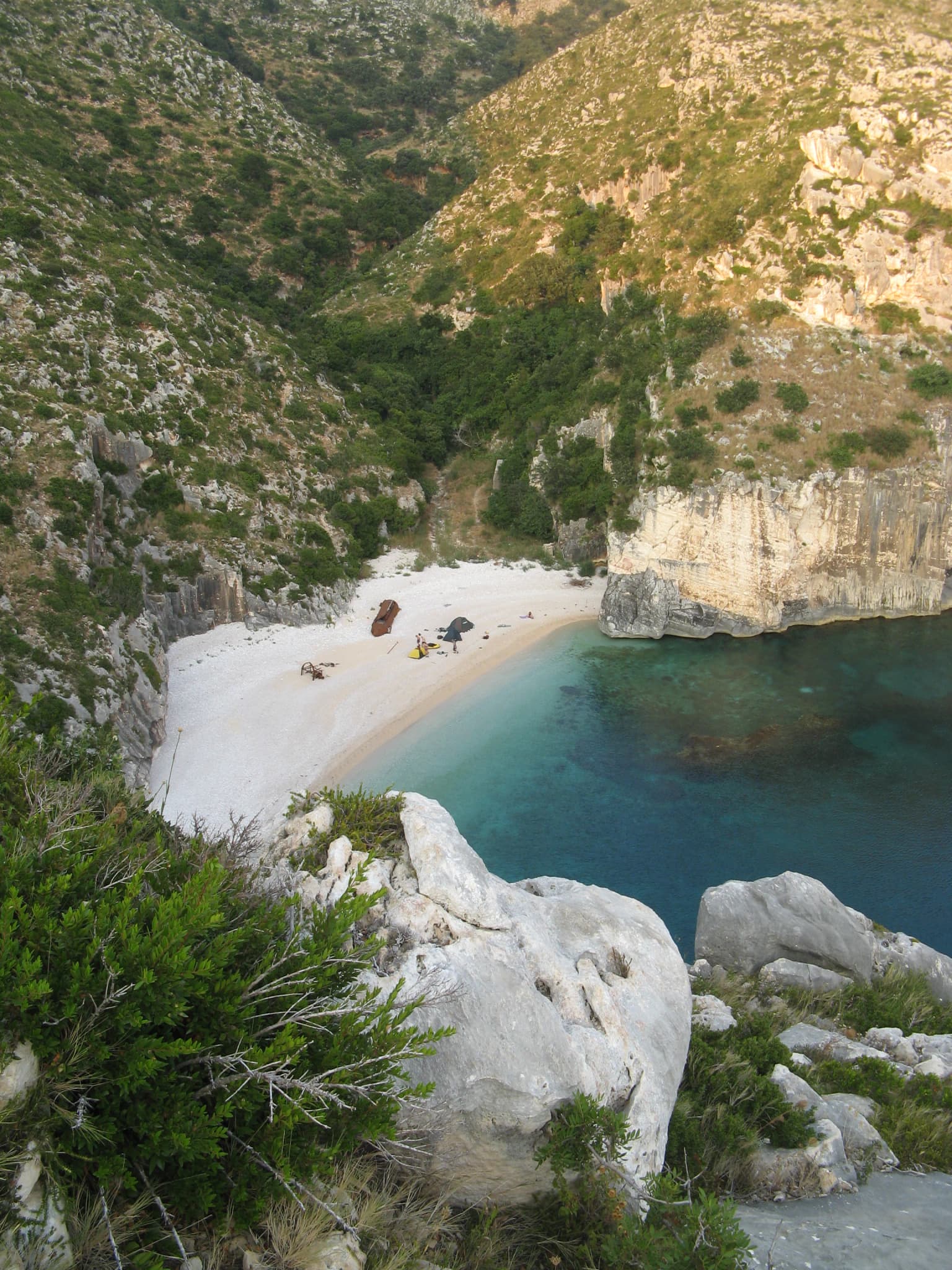 A coastal bay with white sand beach, turquoise water, rocky cliffs, and green vegetation viewed from an elevated vantage point