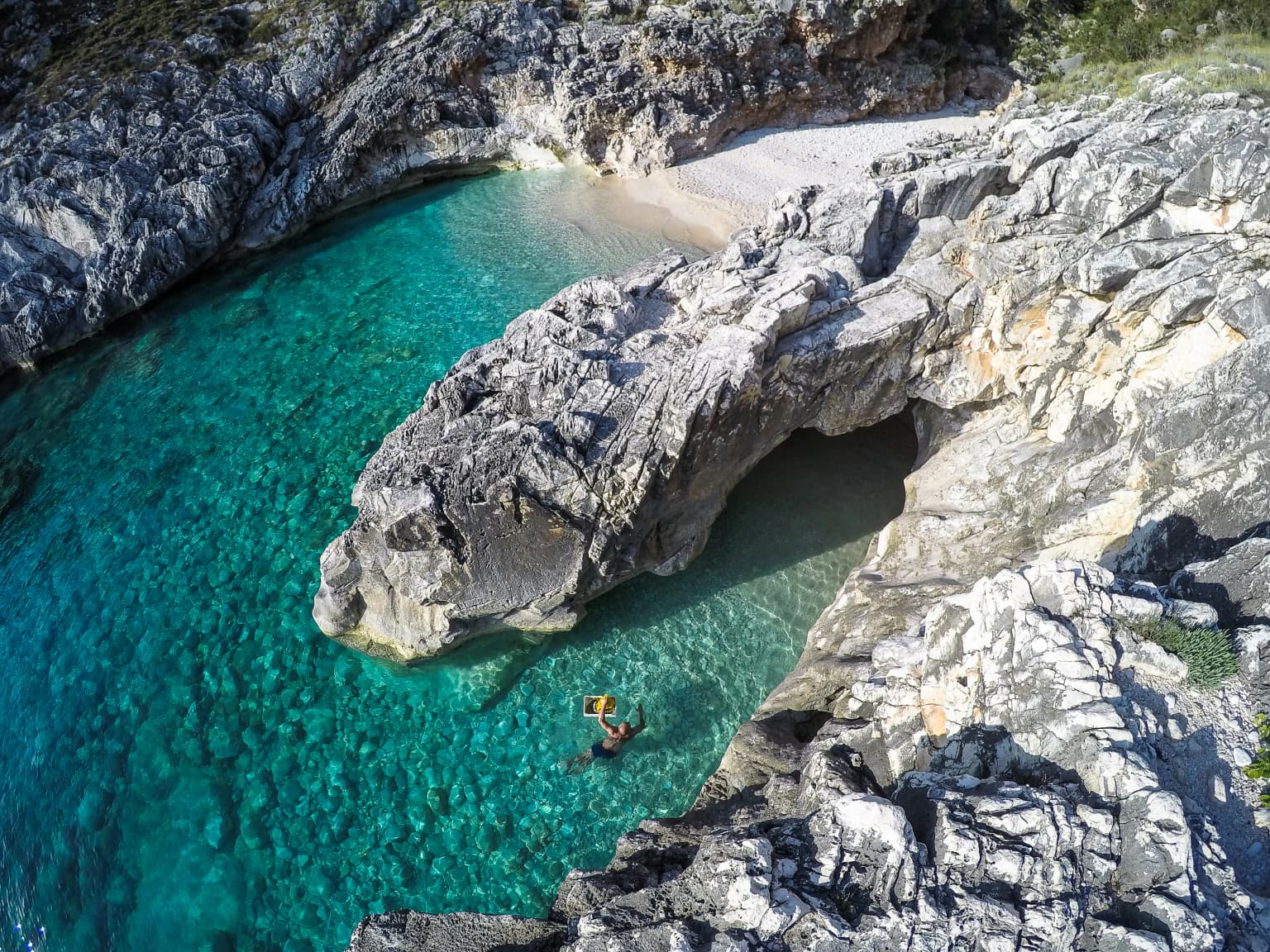 Aerial view of a rocky coastal cove with clear turquoise water, a small sandy beach, and a person swimming near the shore