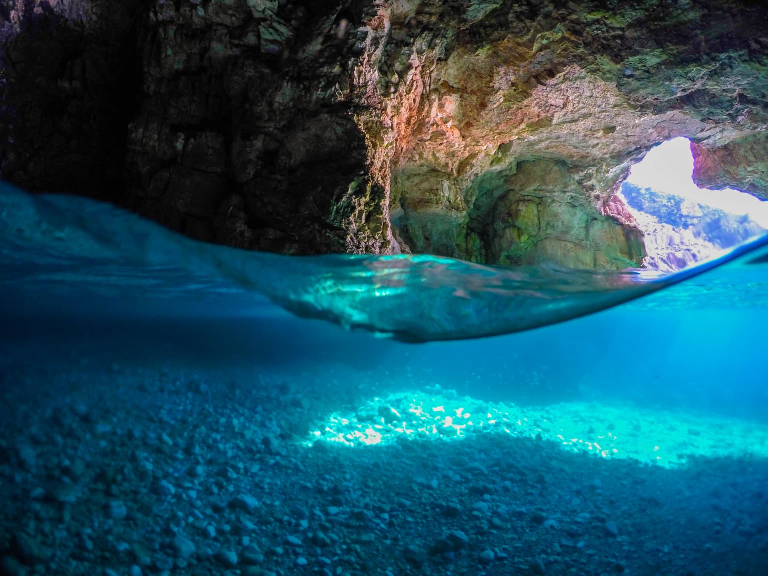 Underwater scene showing cave entrance with rocky walls, clear blue water, and sunlit seabed