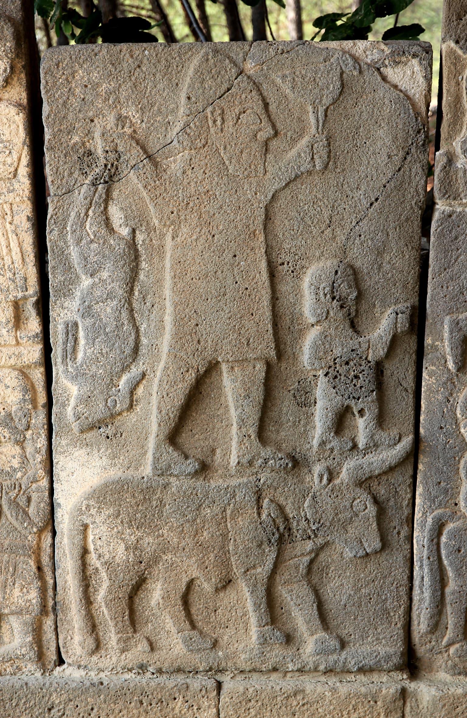Stone relief carving showing a standing figure holding a staff, a smaller figure, a bull, and a mythical creature on a stone slab