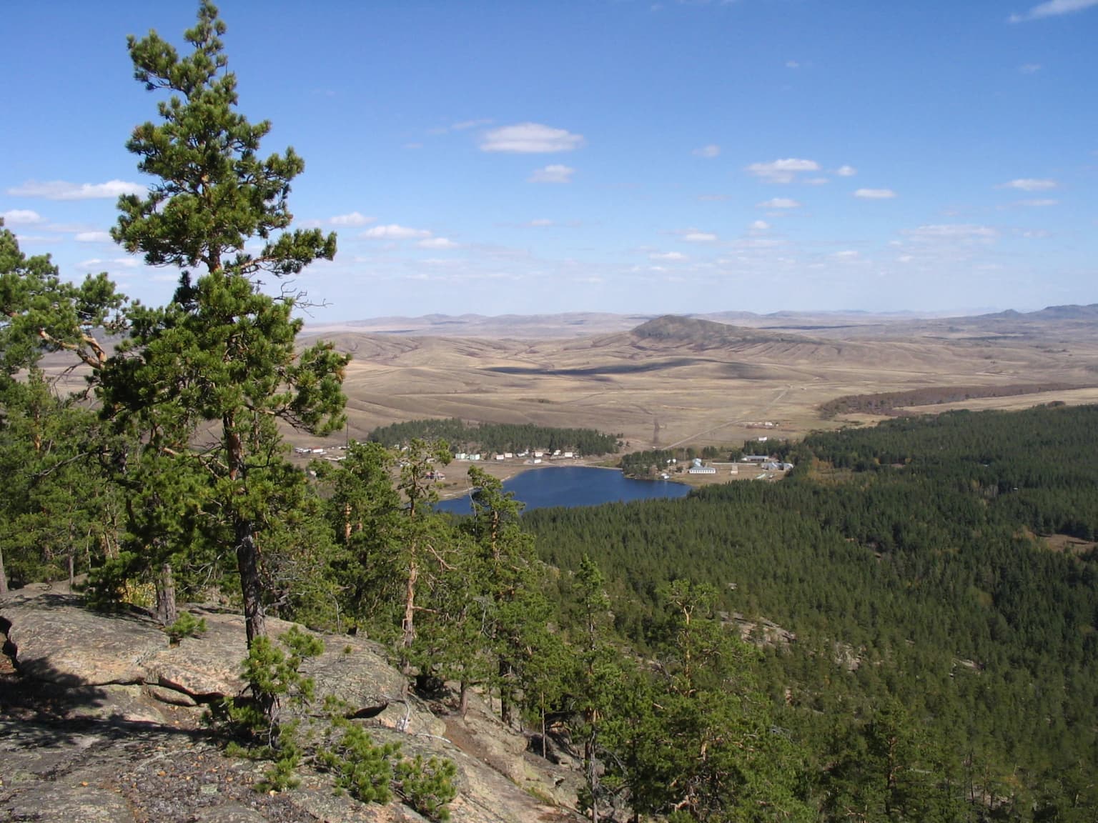 Rocky outcrop with pine trees overlooking a lake surrounded by dense forests and rolling hills under a clear blue sky