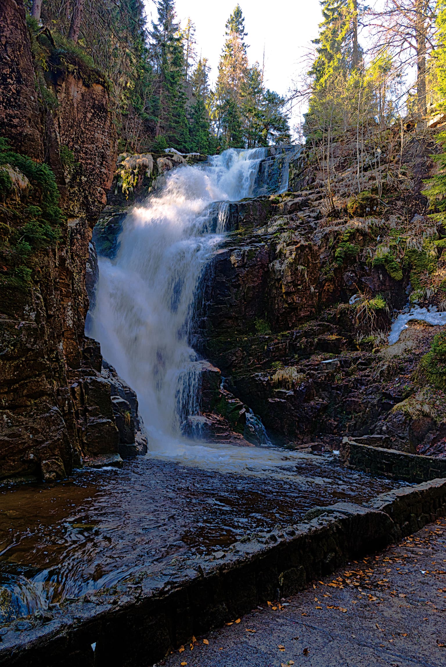 Waterfall flowing over rocky cliffs surrounded by forest with stone pathway in foreground