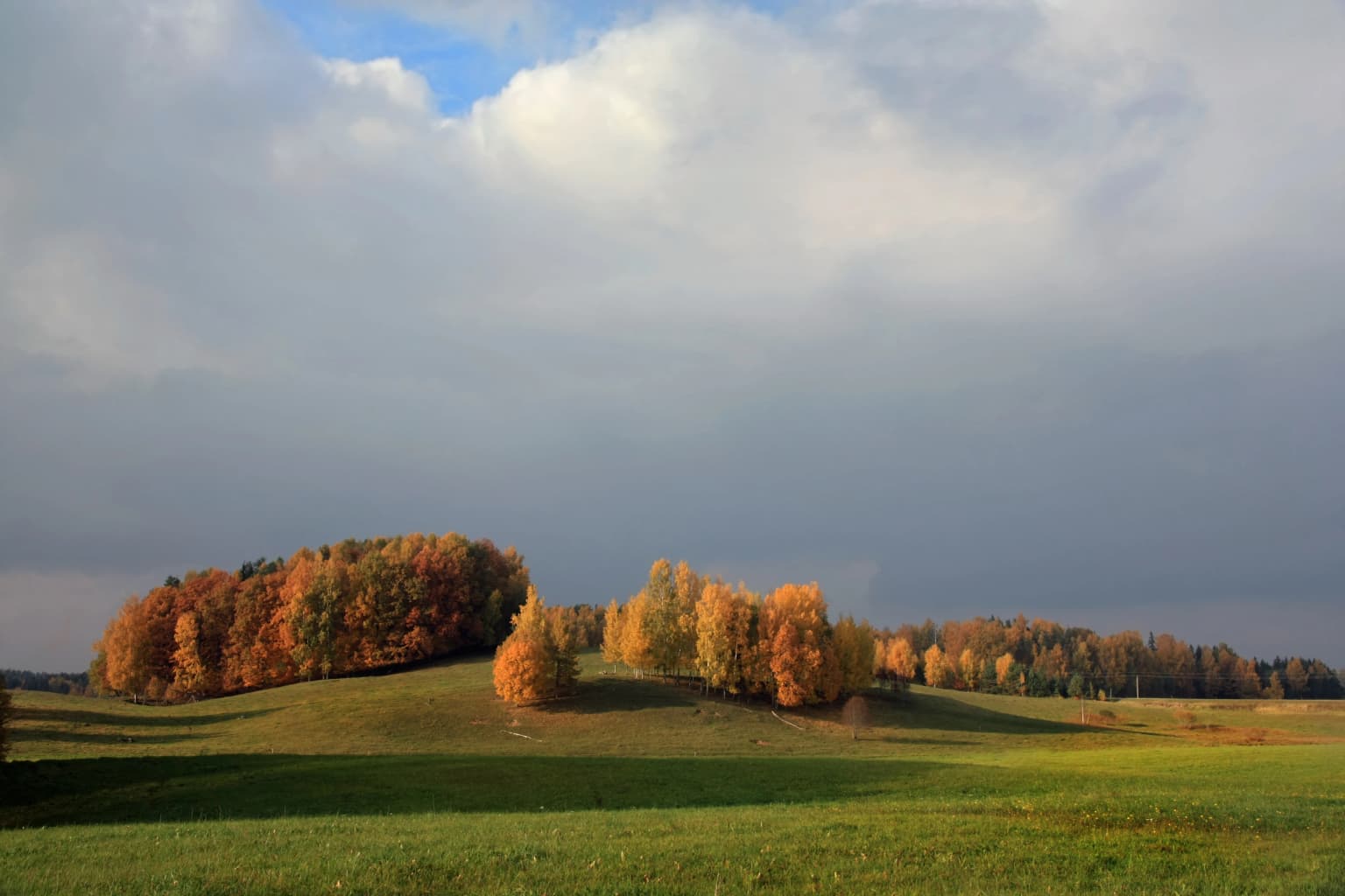 Rolling green fields and hills with autumn-colored trees under a partly cloudy sky