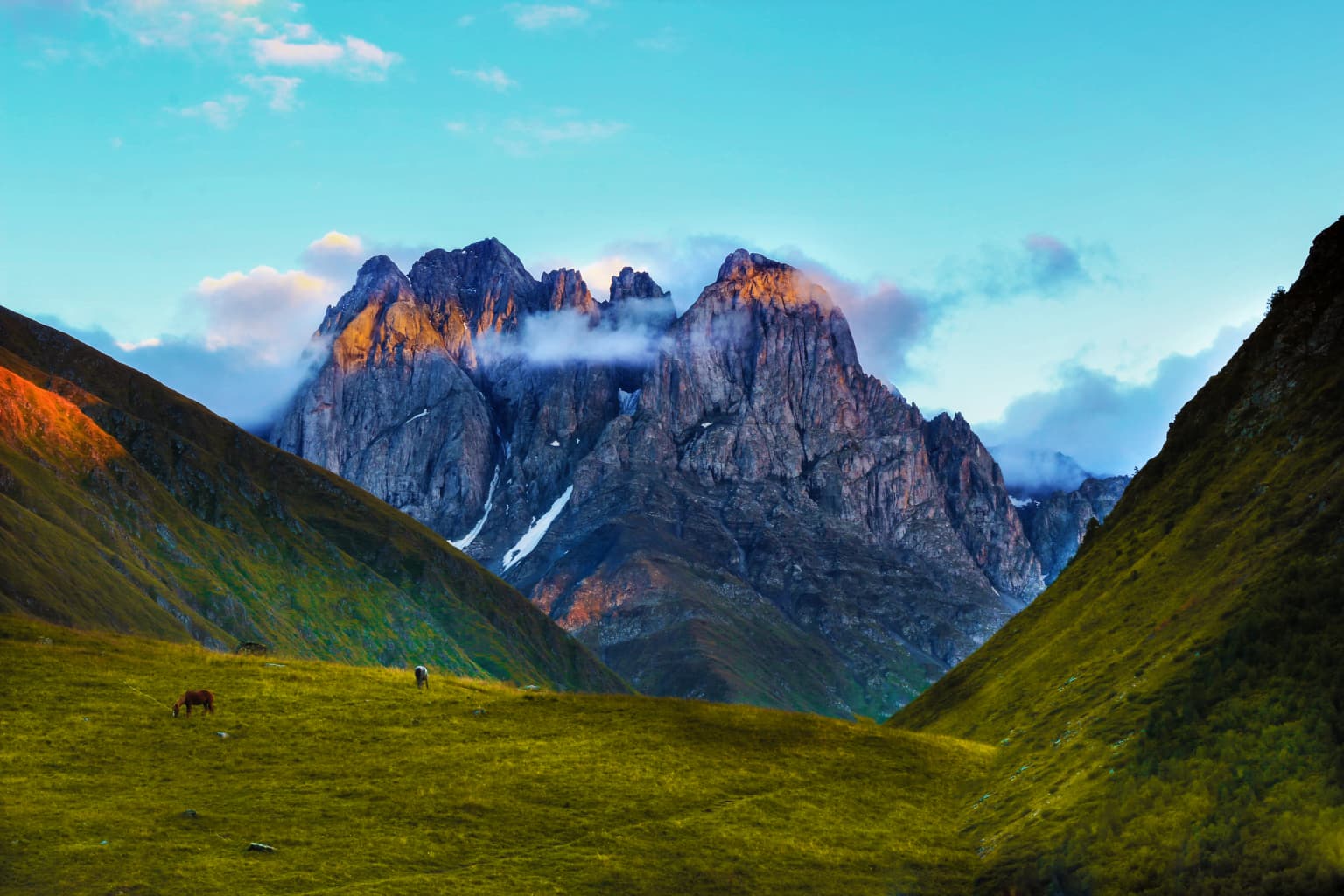 Grassy field with a single animal grazing, surrounded by steep green mountains leading to rocky peaks with patches of snow, under a partly cloudy blue sky