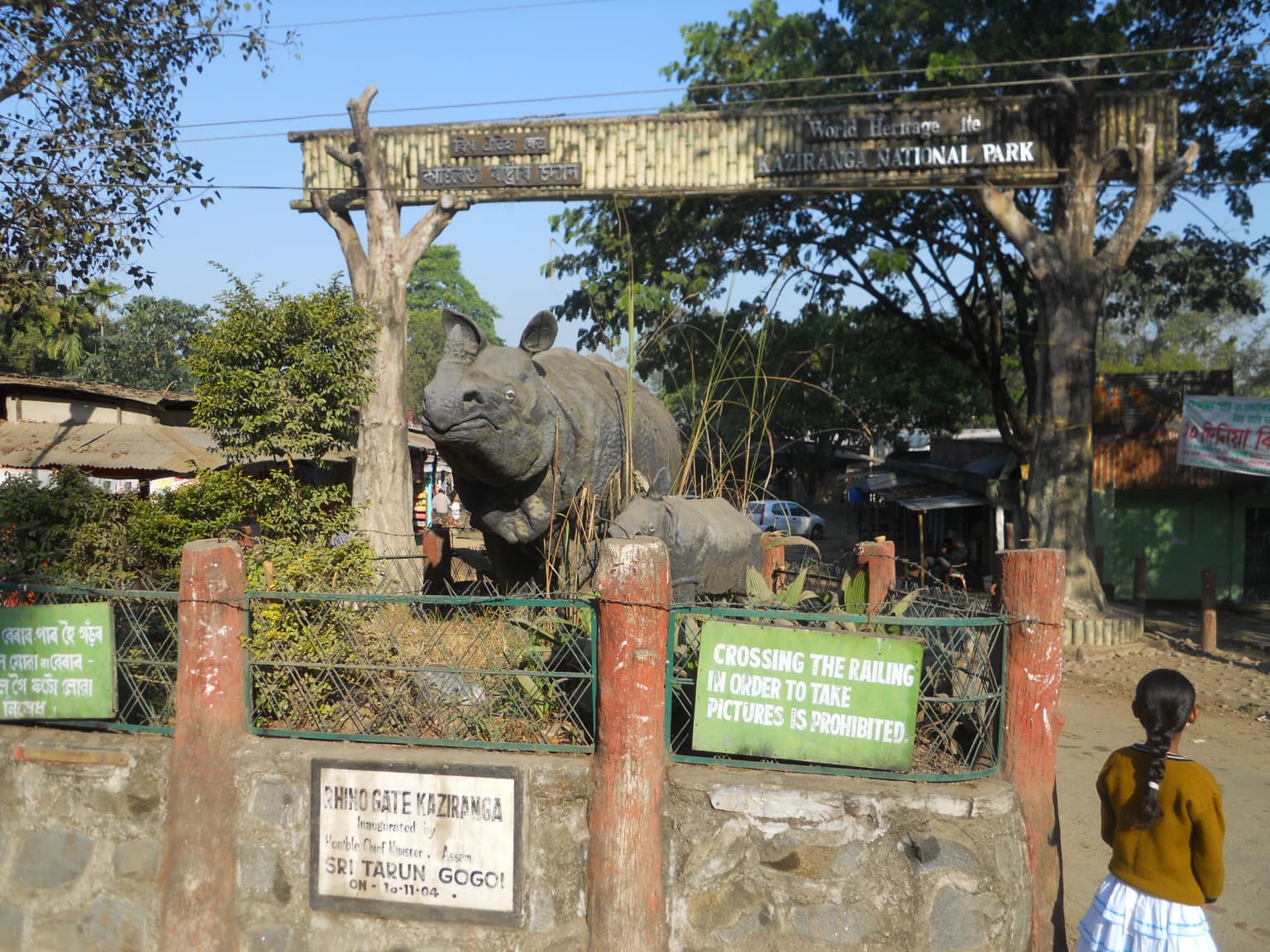 Stone wall entrance gate with red pillars, green signs, and a large rhinoceros statue at Kaziranga National Park. A person stands near the gate.