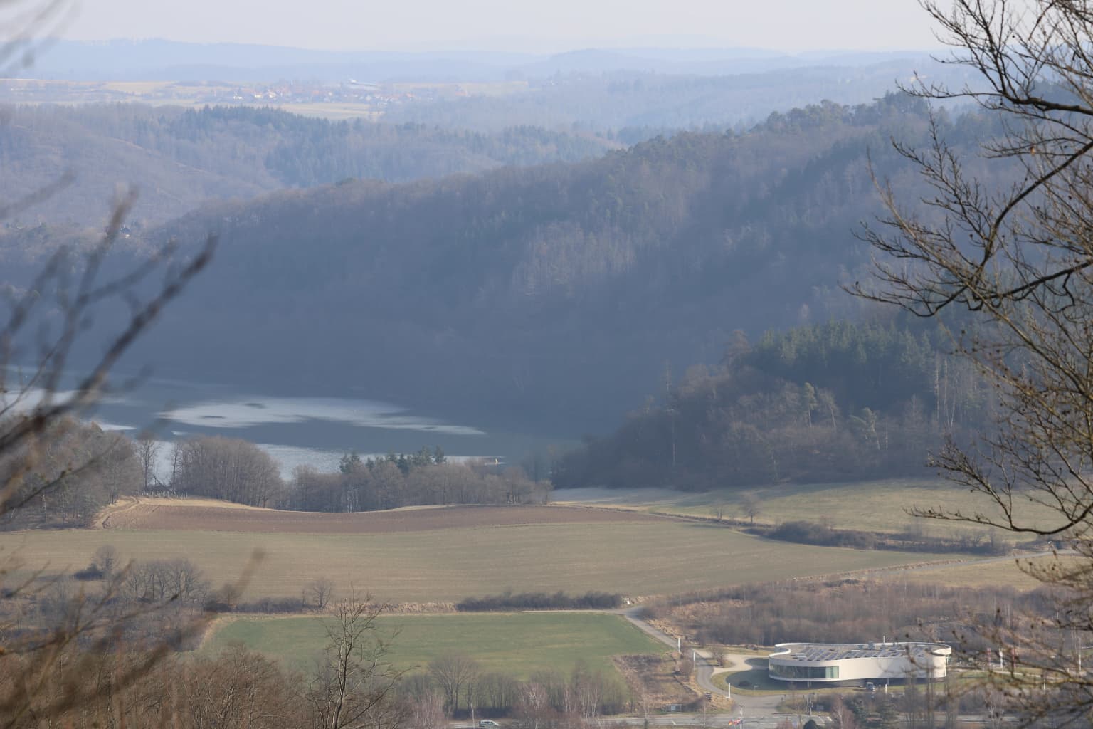 Wide landscape view showing Edersee lake, rolling hills, forested areas, and open fields with a building in the foreground