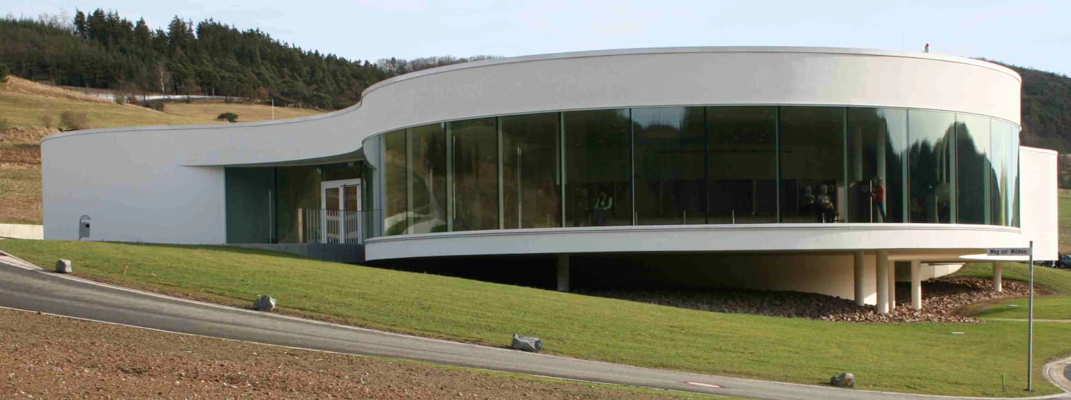 Modern white building with large glass windows on a grassy hillside near a road, with hills in the background
