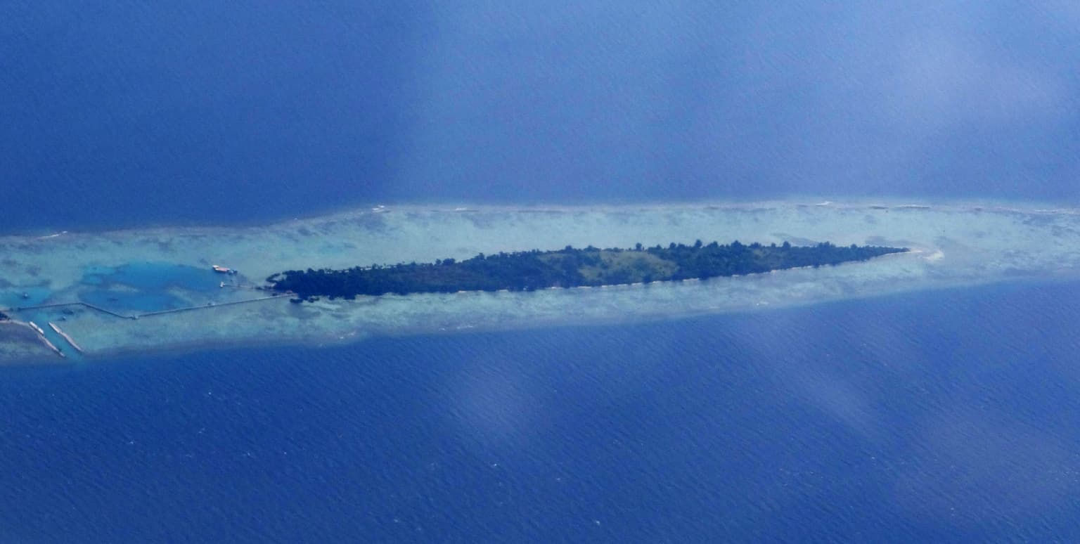 Aerial view of a long narrow island surrounded by blue water with visible coral reef