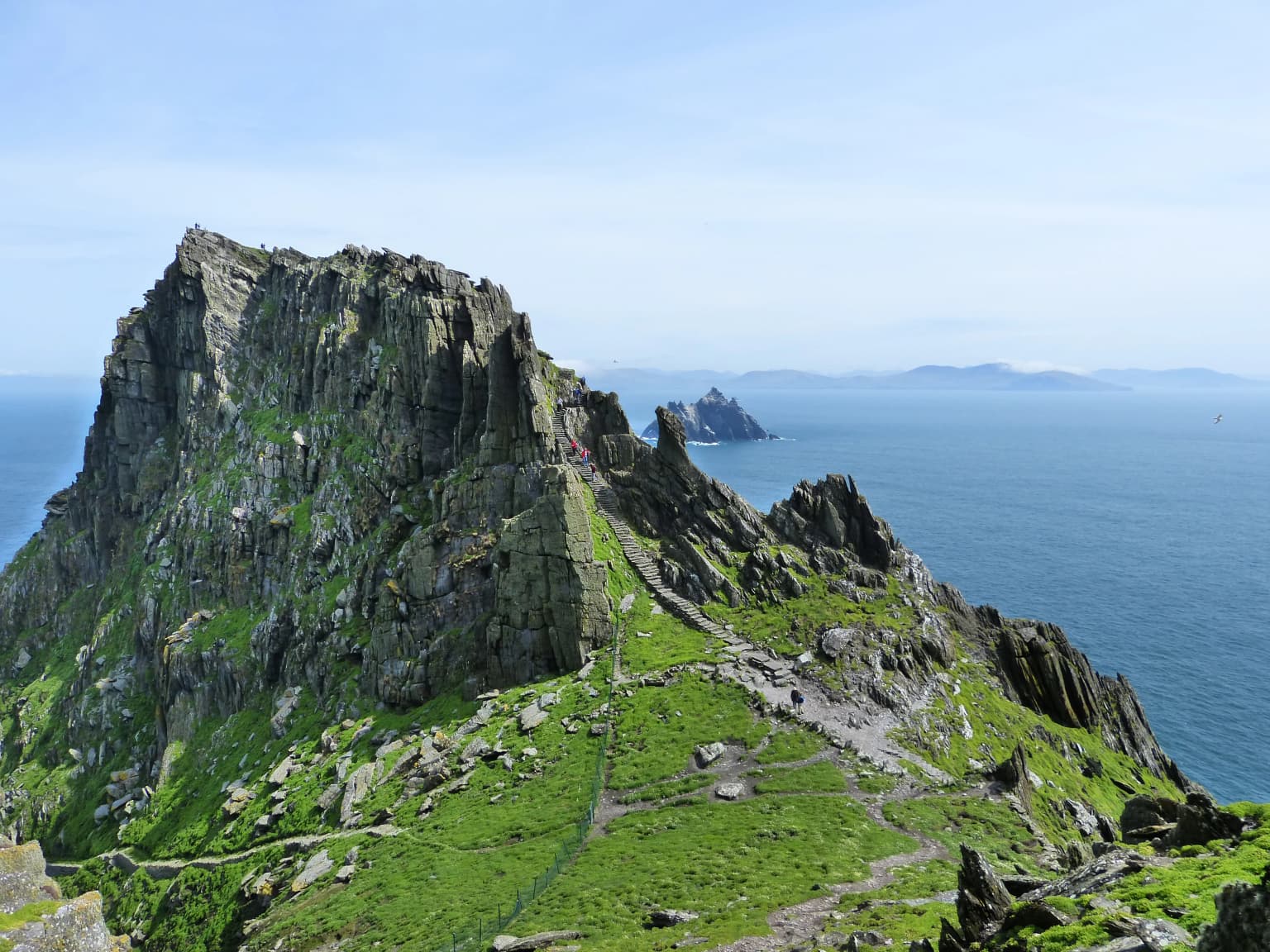 Rugged rocky island with green vegetation and a narrow path leading to the summit, with ocean and distant islands in the background