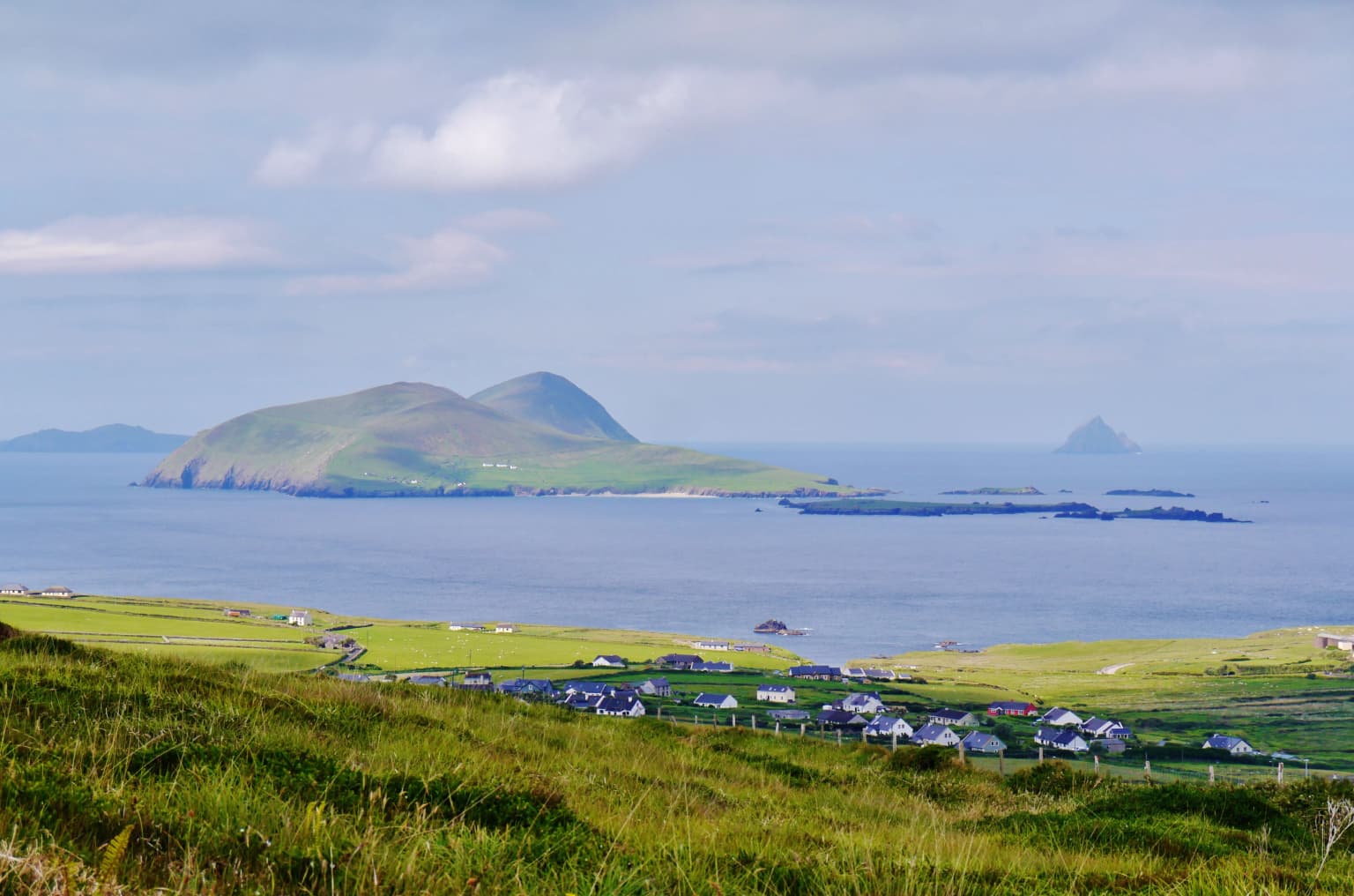 Coastal landscape with green fields, scattered buildings, a large island in the sea, and smaller islands in the distance under a partly cloudy sky