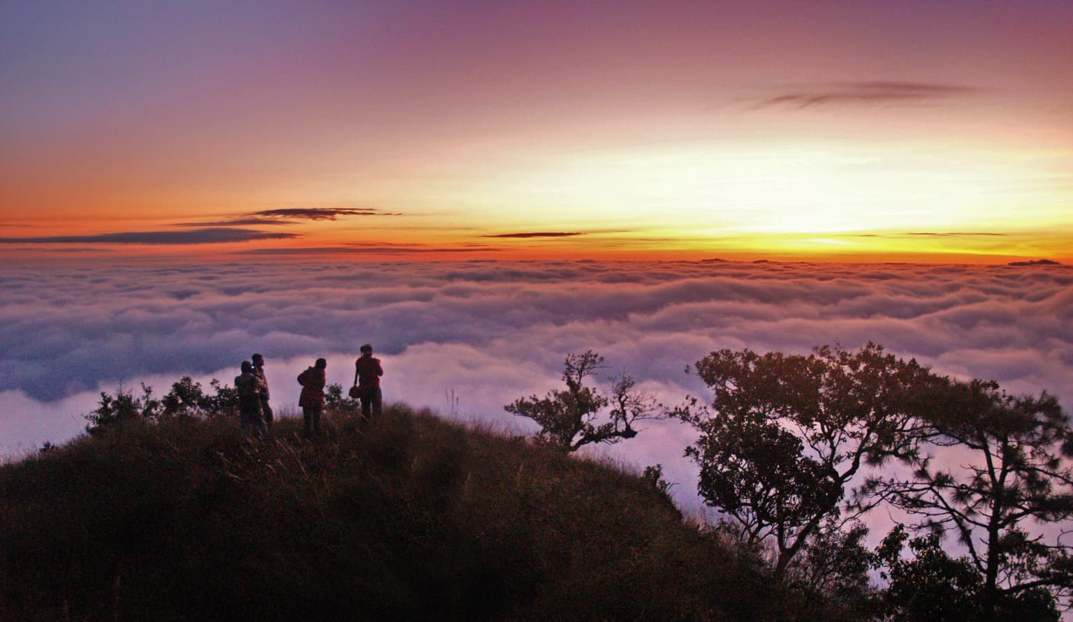 Grupo de excursionistas de pie en la cima de una montaña con nubes debajo durante el amanecer