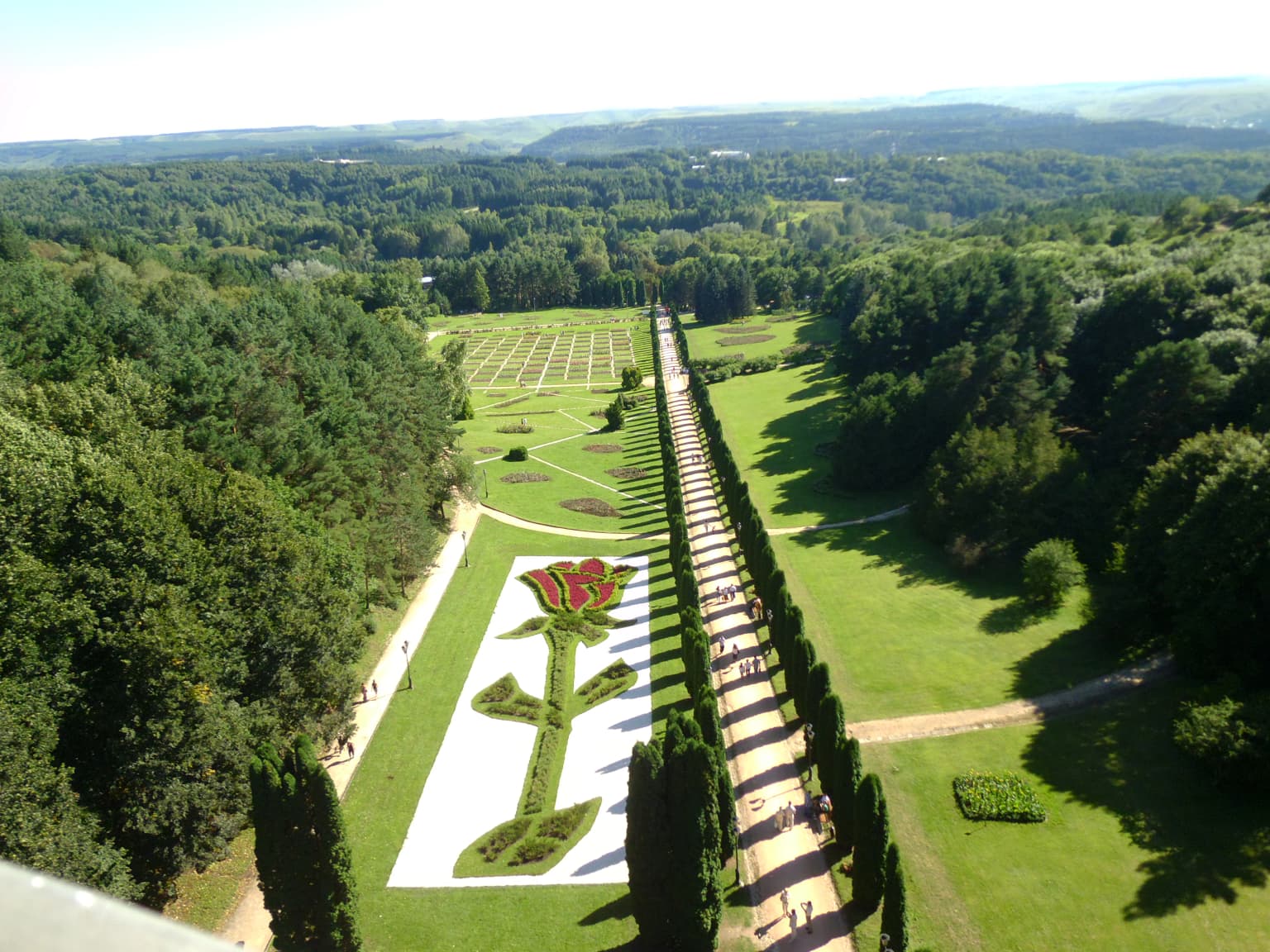 Aerial view showing a large garden with a rose-shaped floral design, surrounded by green fields, trees, and pathways with people walking