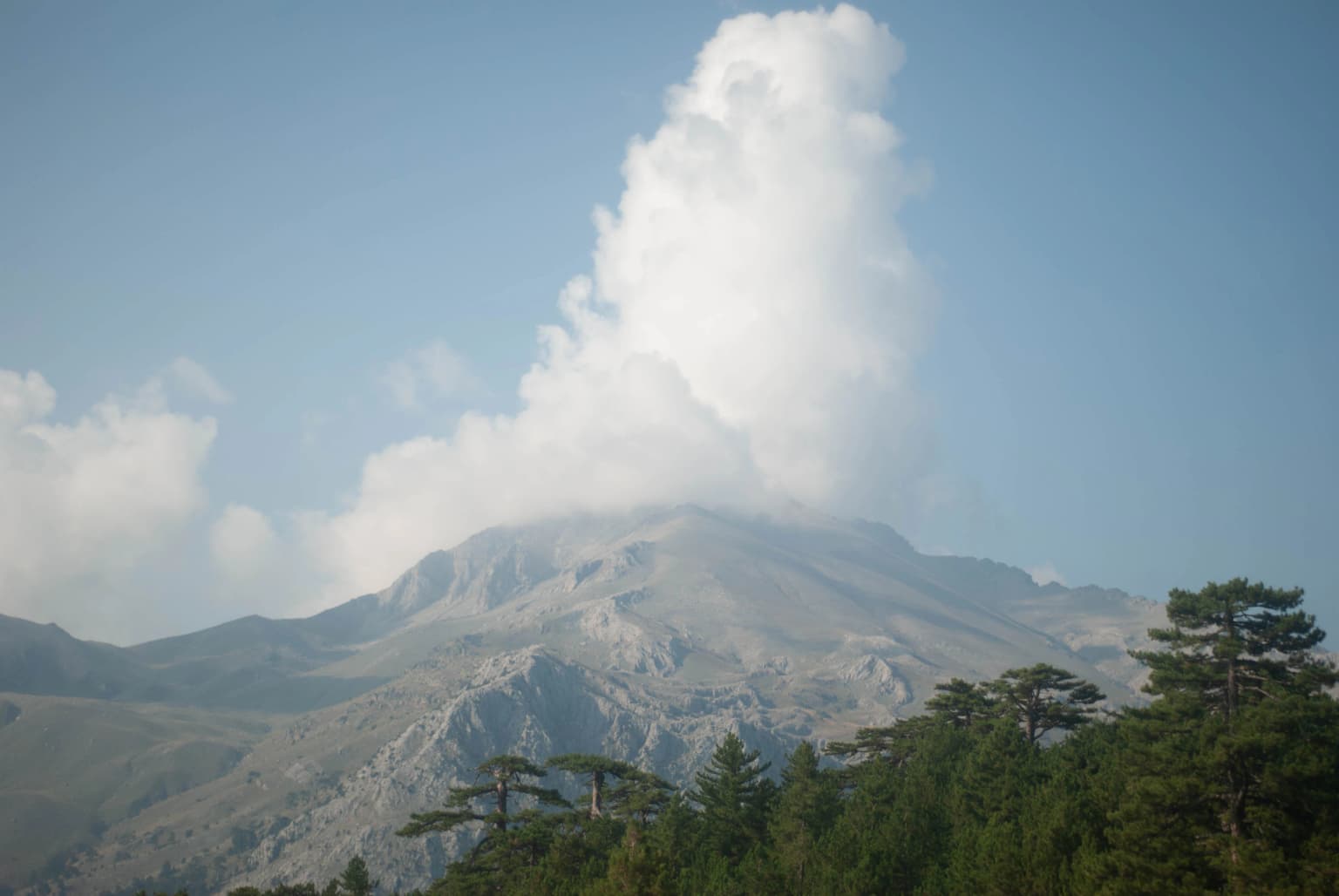 Mountain peak with clouds, forested foreground, and clear blue sky