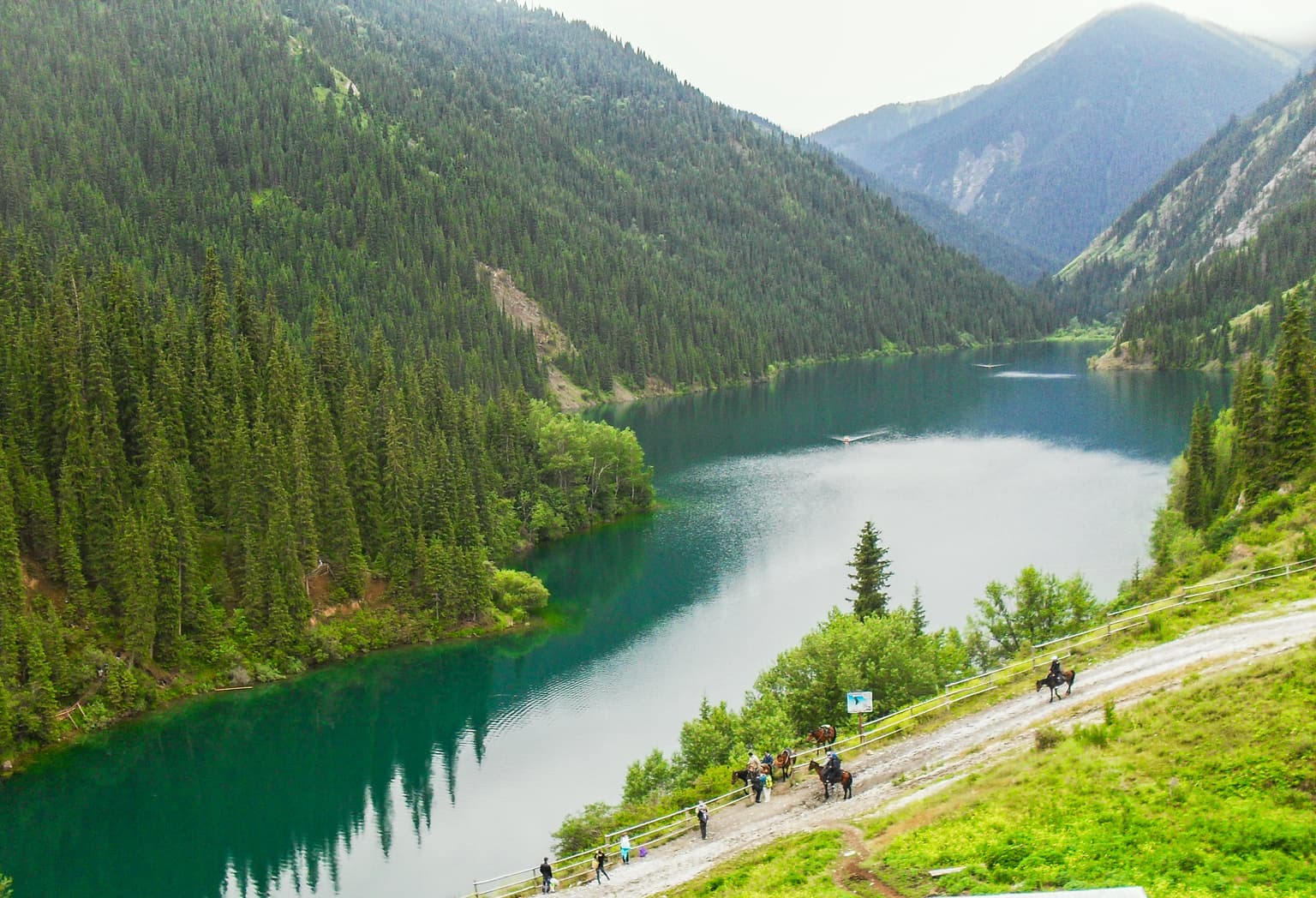 Panoramic view of Kolsay Lake with vibrant turquoise water, flanked by lush green forests and towering mountains, with a road and people visible on the right