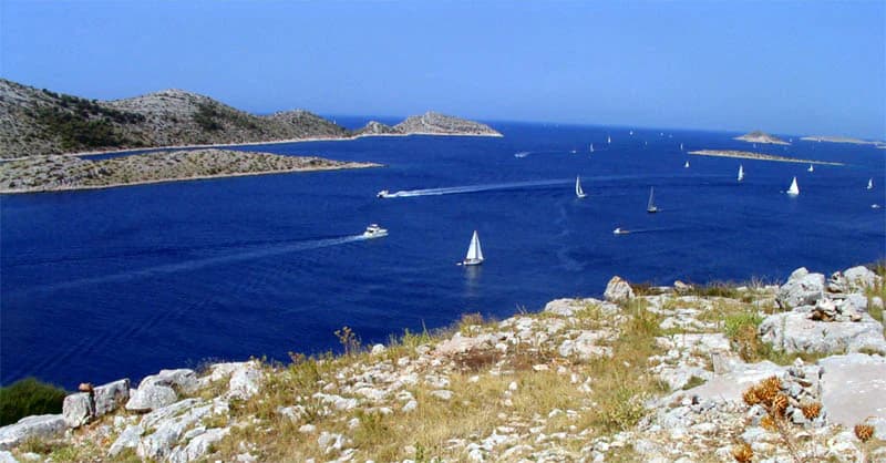 Rocky shoreline with dry grass, blue sea, multiple sailboats, and distant islands under clear blue sky