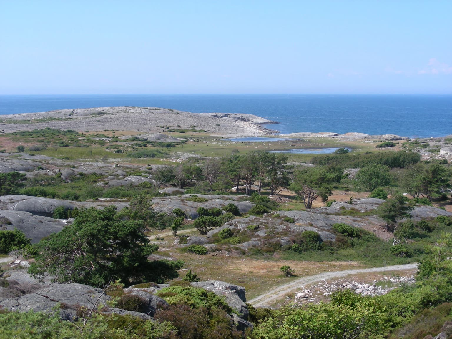 Rocky terrain with scattered vegetation, a winding path, and the sea visible under a clear blue sky
