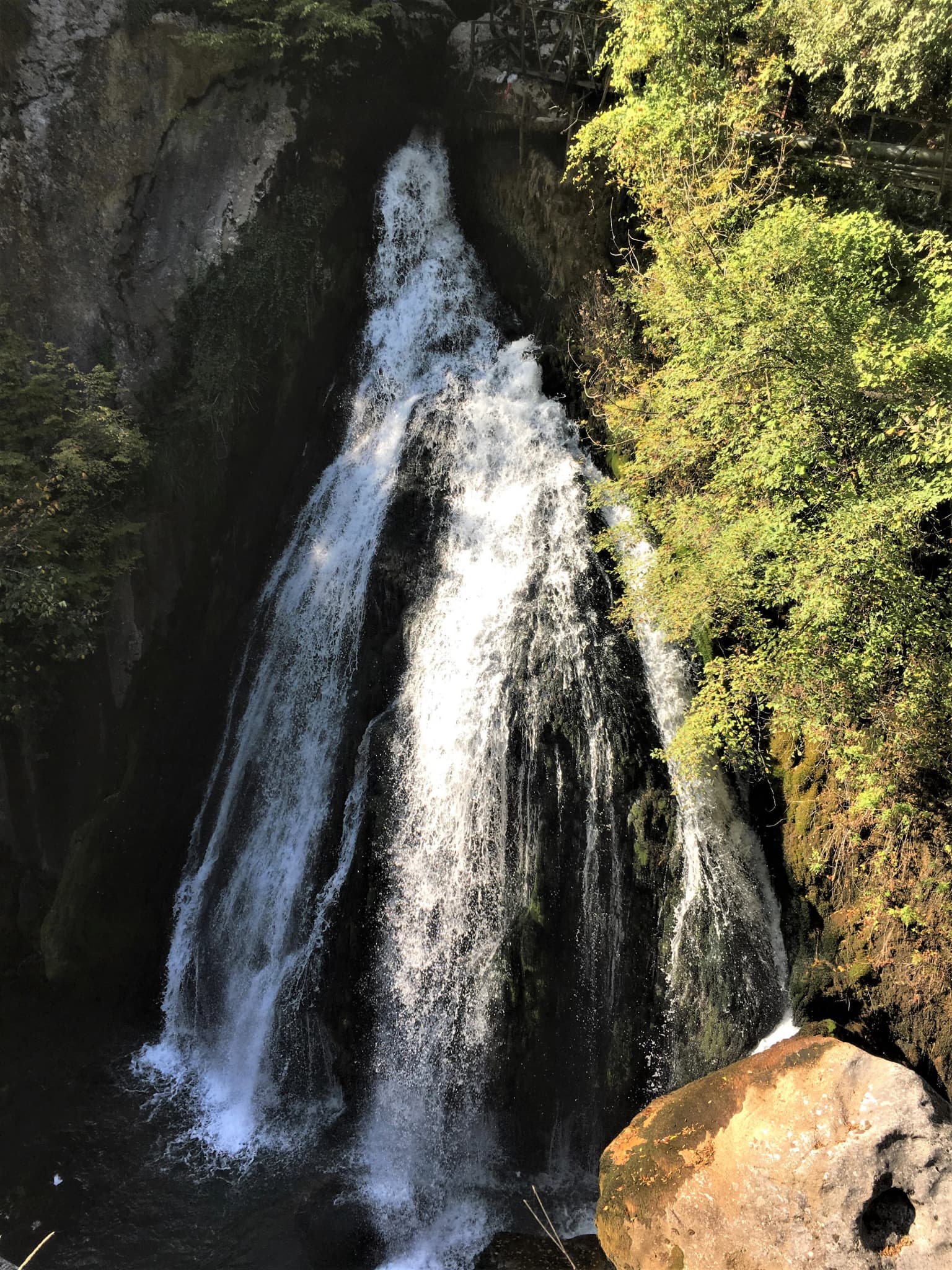 Waterfall cascading down rocky cliffs with dense green foliage on the right side and a large boulder in the foreground