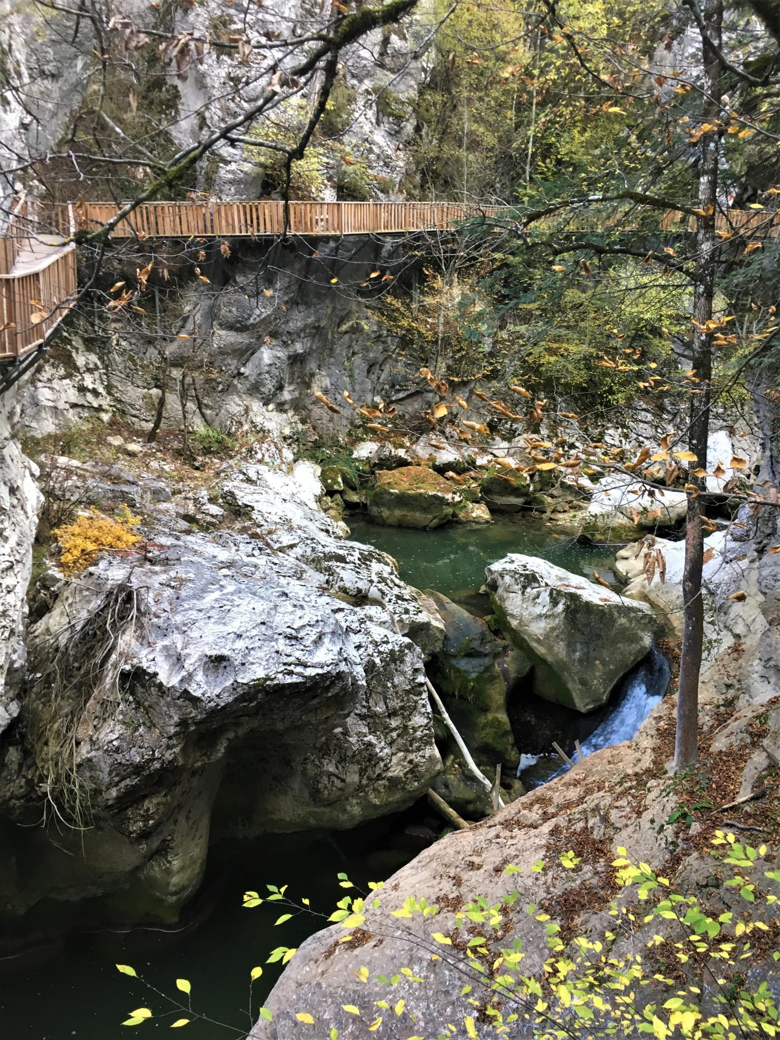 Small waterfall cascading over rocks in a canyon with a wooden bridge spanning the gorge, surrounded by trees and autumn foliage
