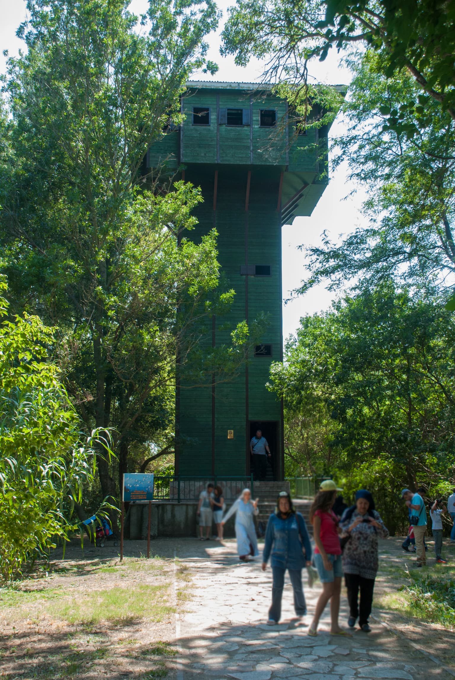 Tall green observation tower surrounded by trees with several people walking on a stone pathway
