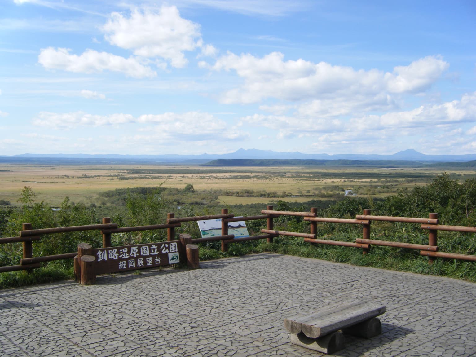 Plataforma de observación de madera con señalización japonesa que domina un vasto paisaje de humedales con montañas en la distancia bajo un cielo parcialmente nublado