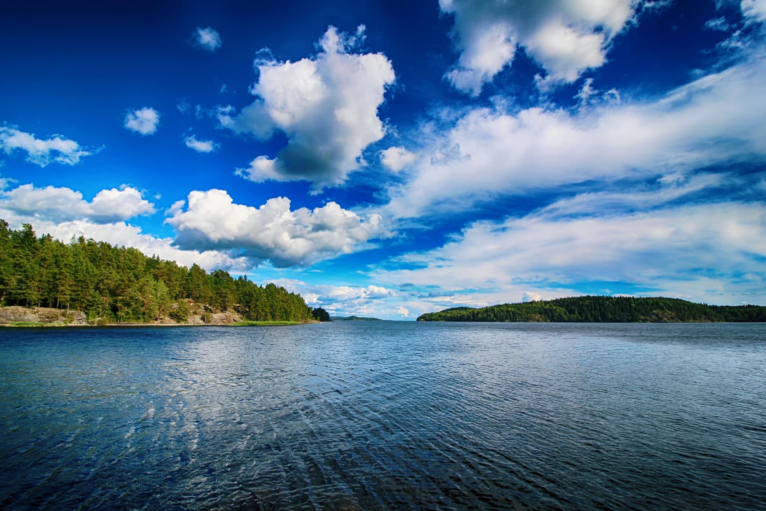 Calm lake water with forested islands in the distance under a blue sky with scattered clouds