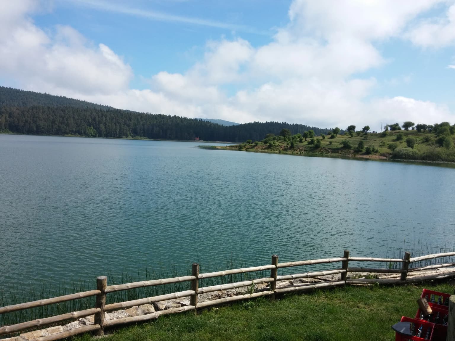 Lake Abant with wooden fence in foreground, grassy shore, and forested hills under partly cloudy sky