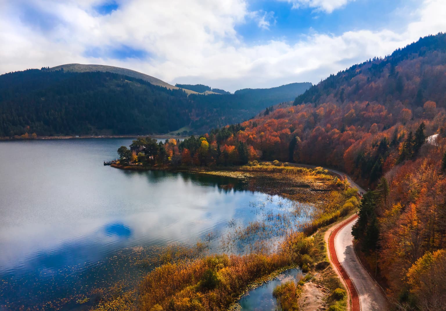 Aerial view of a lake surrounded by autumn-colored forests and hills with a road along the shore
