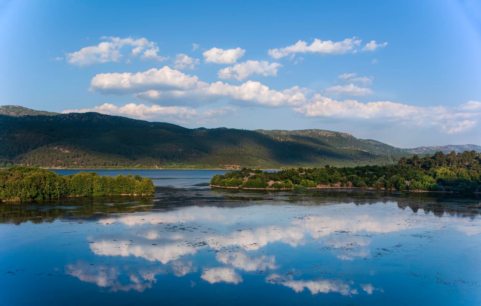 Lake Kovada reflecting clouds and sky with mountains in the background.