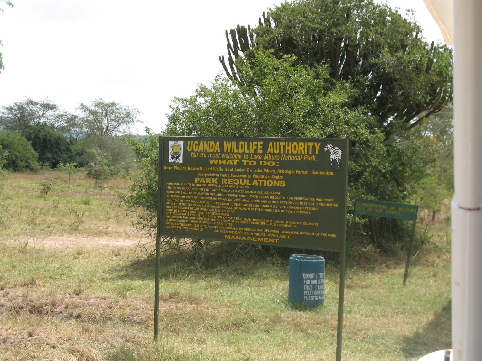 Green sign displaying Uganda Wildlife Authority and park regulations at Lake Mburo National Park entrance with grassy field and trees in background